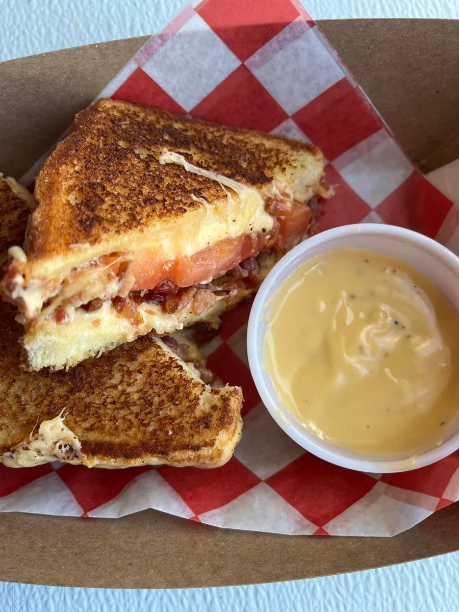 A grilled tomato and cheese sandwich on red-checkered paper with a side of dipping sauce from The Sister Shack.